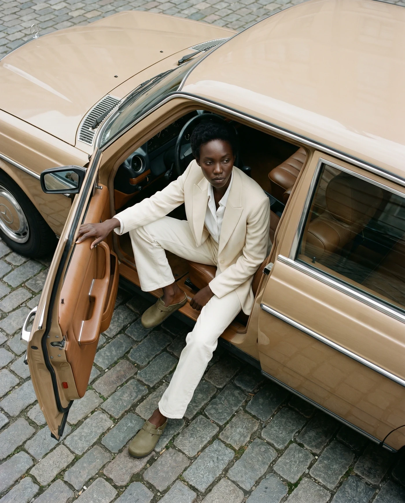AI fashion model in a cream suit stepping out of a vintage sedan on a cobbled street