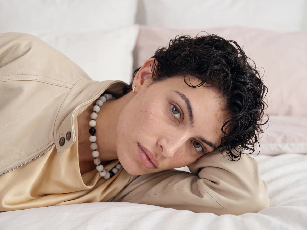 AI brand photoshoot imagery: model resting on bedding in a cream leather jacket and pearl necklace