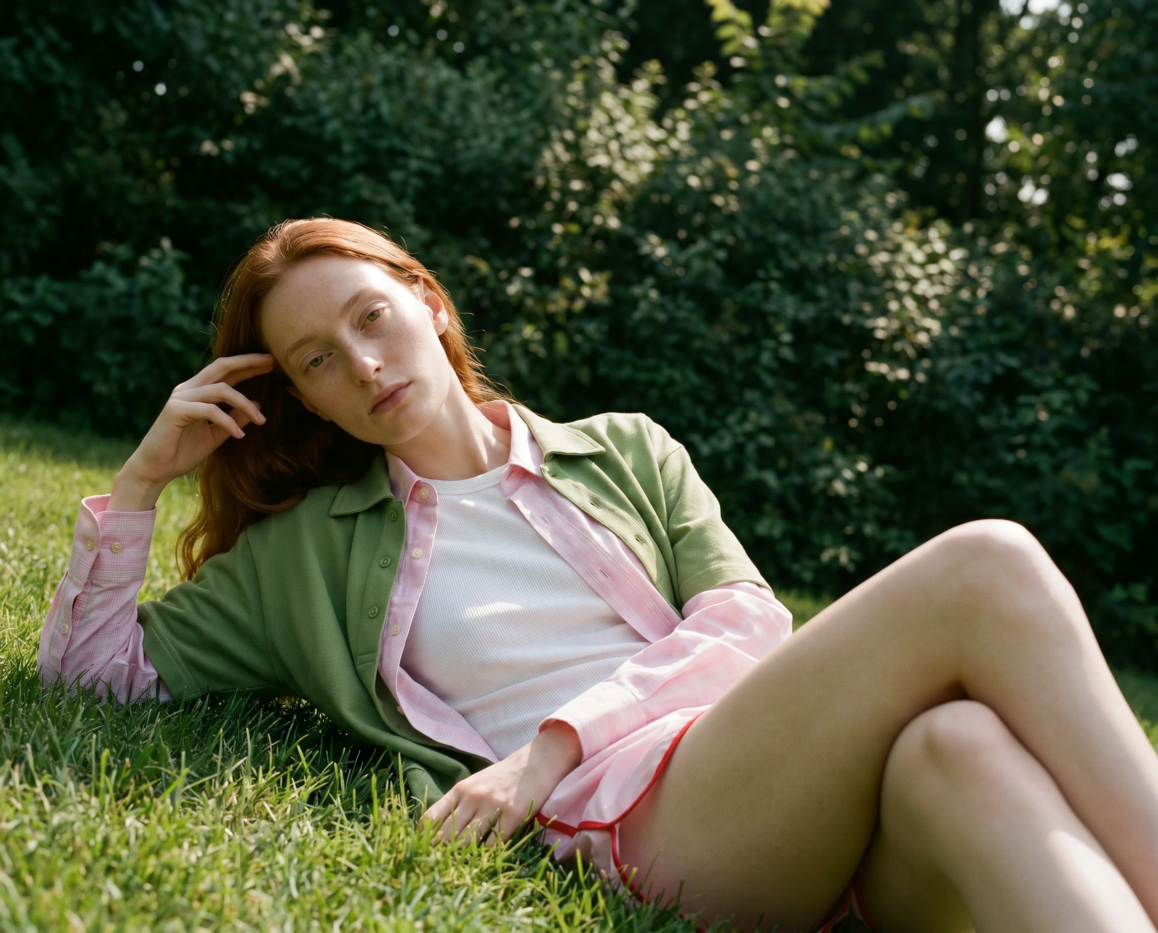 On-model imagery from a ghost mannequin archive: model in a green overshirt layered over a pink check shirt, lying on summer grass