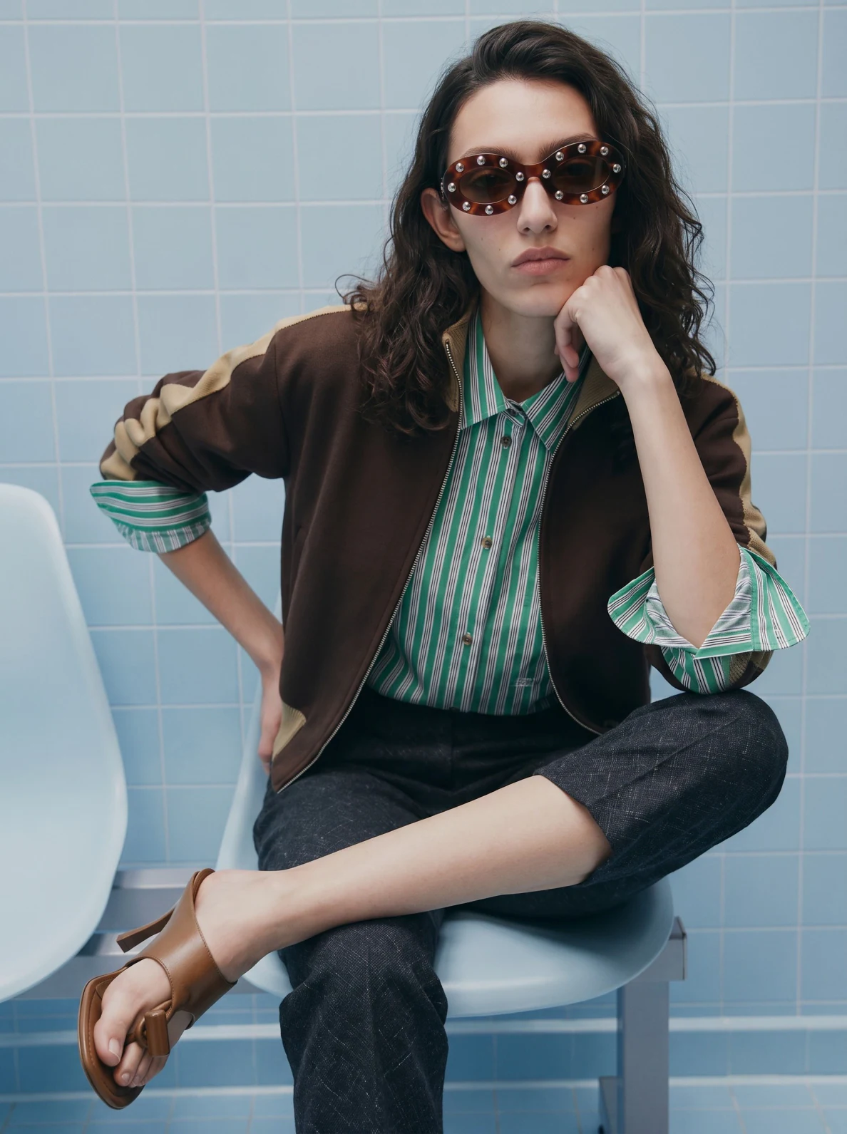 AI-generated fashion image: model in a striped shirt and bomber, seated in a blue-tiled set
